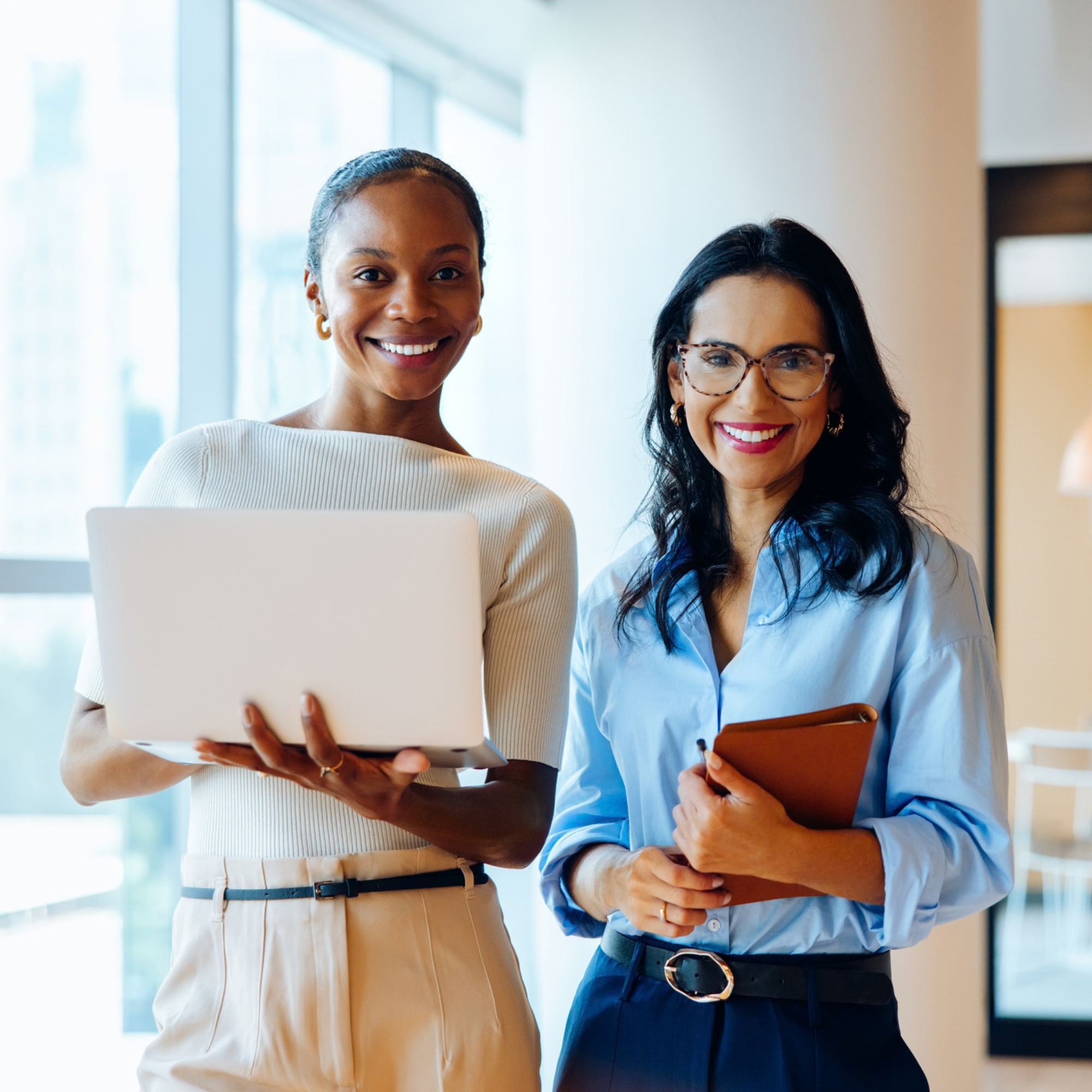 Two women at work, one with a laptop, the other a notebook. Both staring at camera.